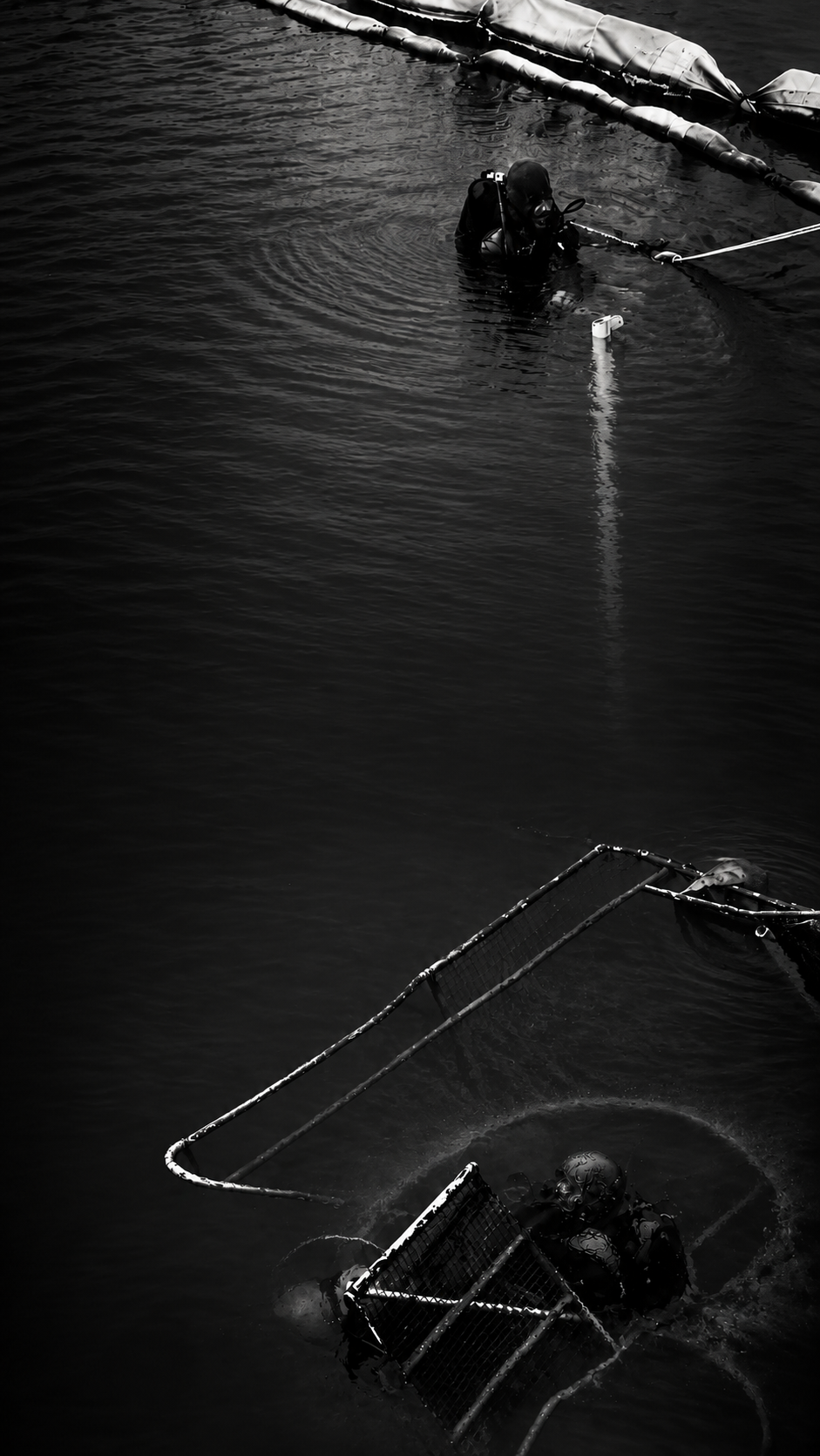 Black-and-white photograph of divers working in dark water beside submerged equipment.