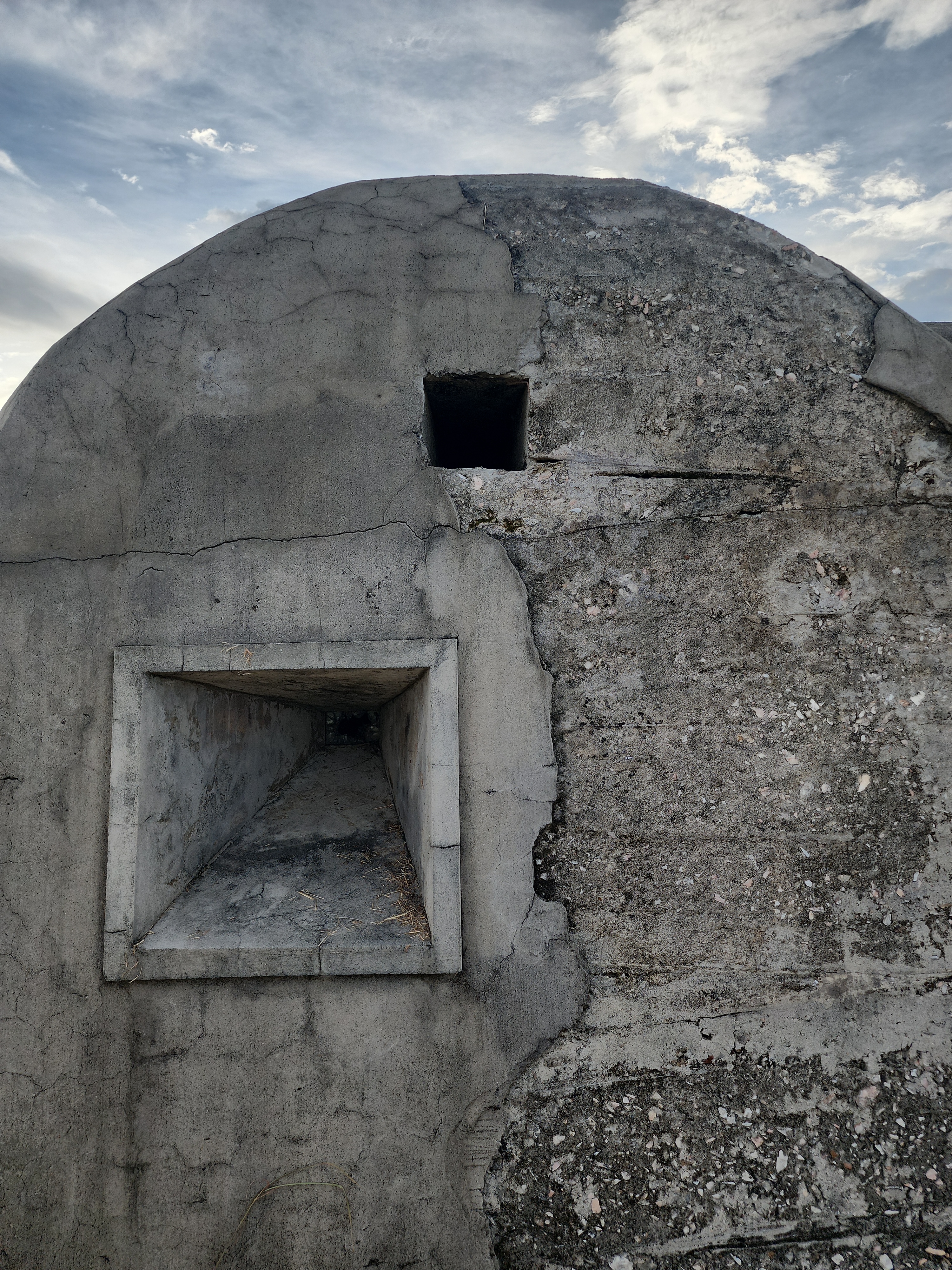 Photograph of the exterior face of a rounded concrete structure with visible cracking, surface loss, a large rectangular opening, and a smaller square opening above it.